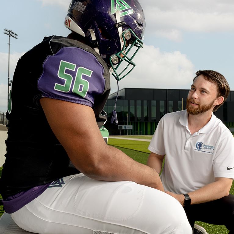 Football player sitting on bench talking to athletic trainer.