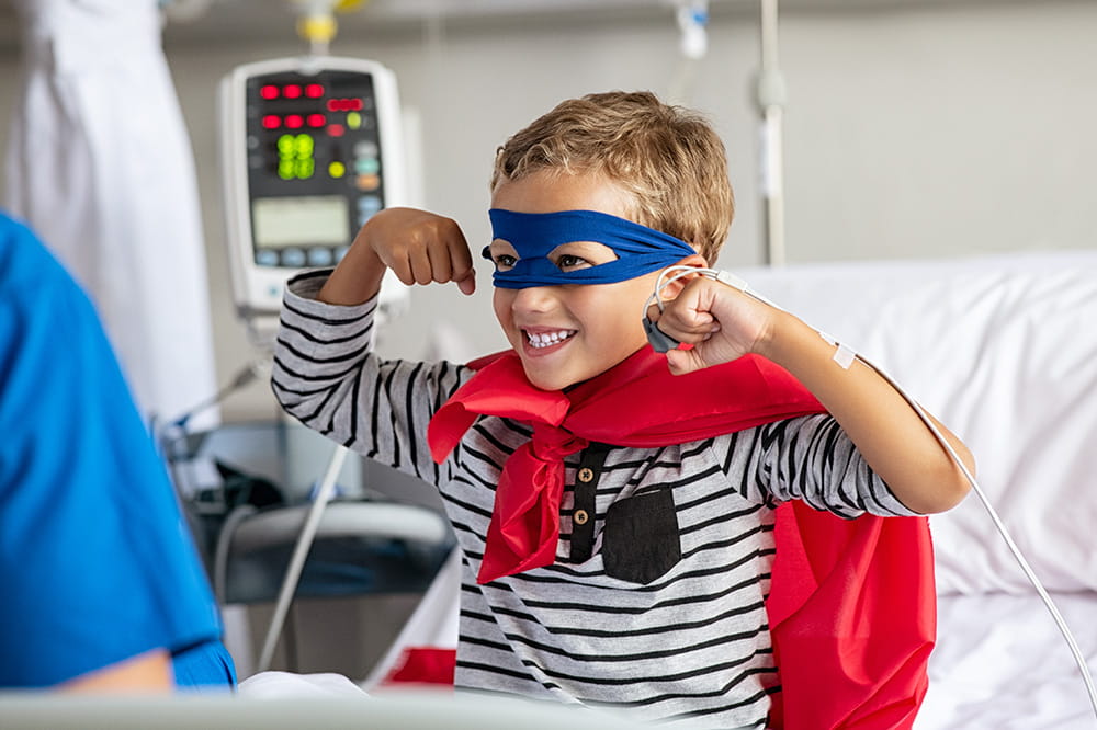 Niño sentado en la cama del hospital con capa y máscara de superhéroe.