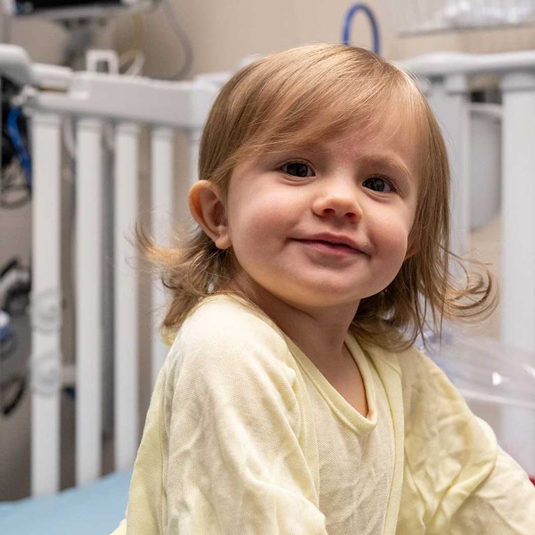Smiling little girl with blonde hair wearing yellow hospital gown.