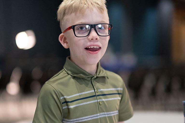 Smiling boy with blonde hair wearing a green shirt and eyeglasses.