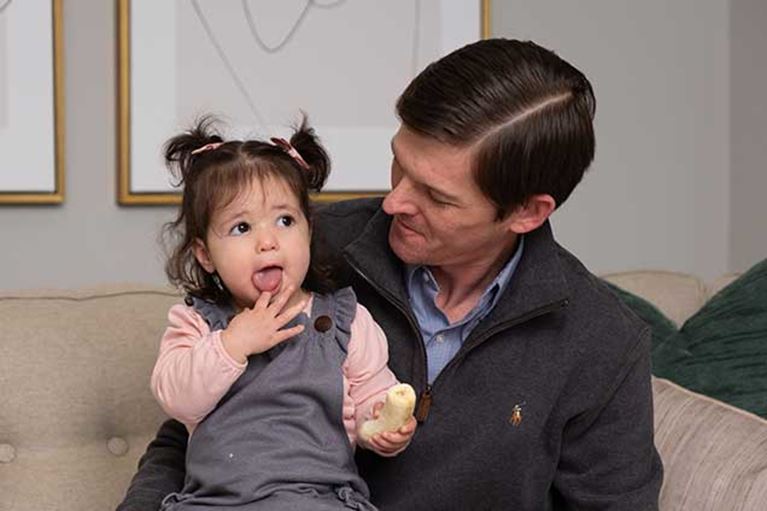 Dad with brown hair sitting on couch holding his daughter that is wearing pigtails in hair. 