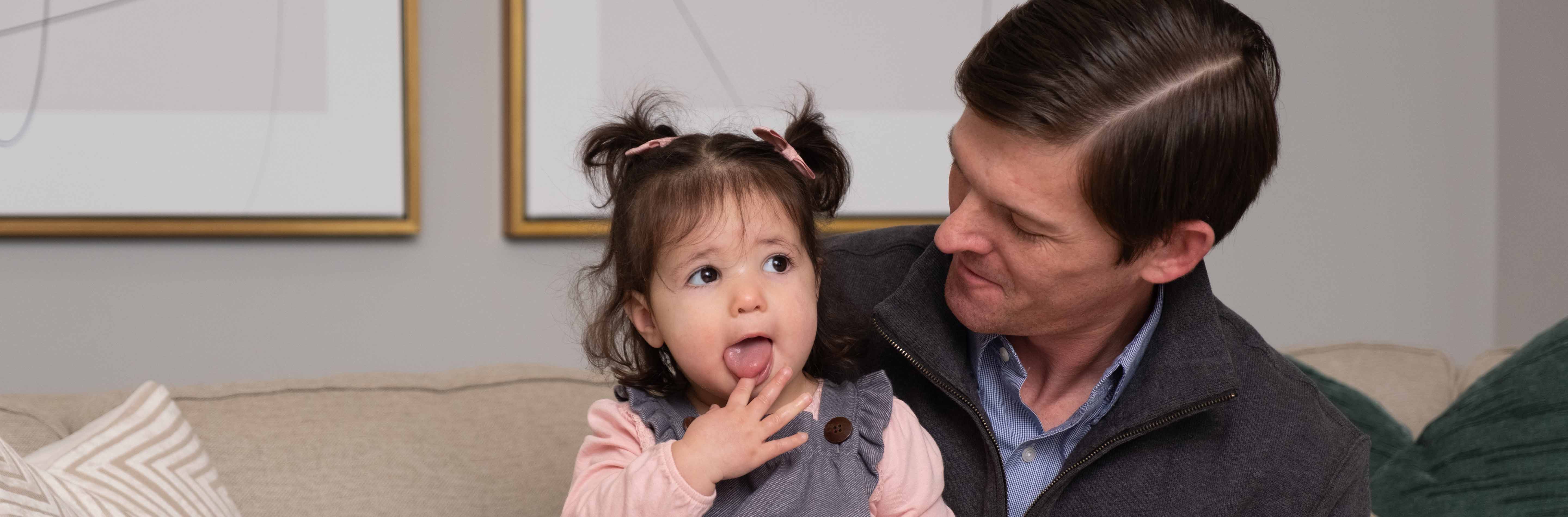 Dad with brown hair sitting on couch holding his daughter that is wearing pigtails in hair. 