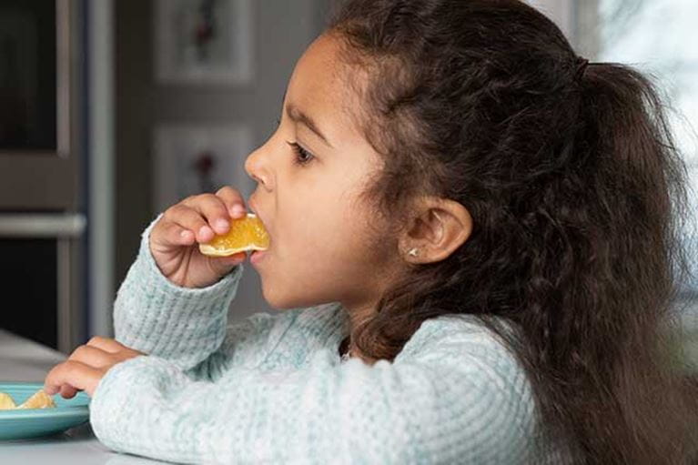 Toddler girl with curly brown hair sitting at table eating an orange.