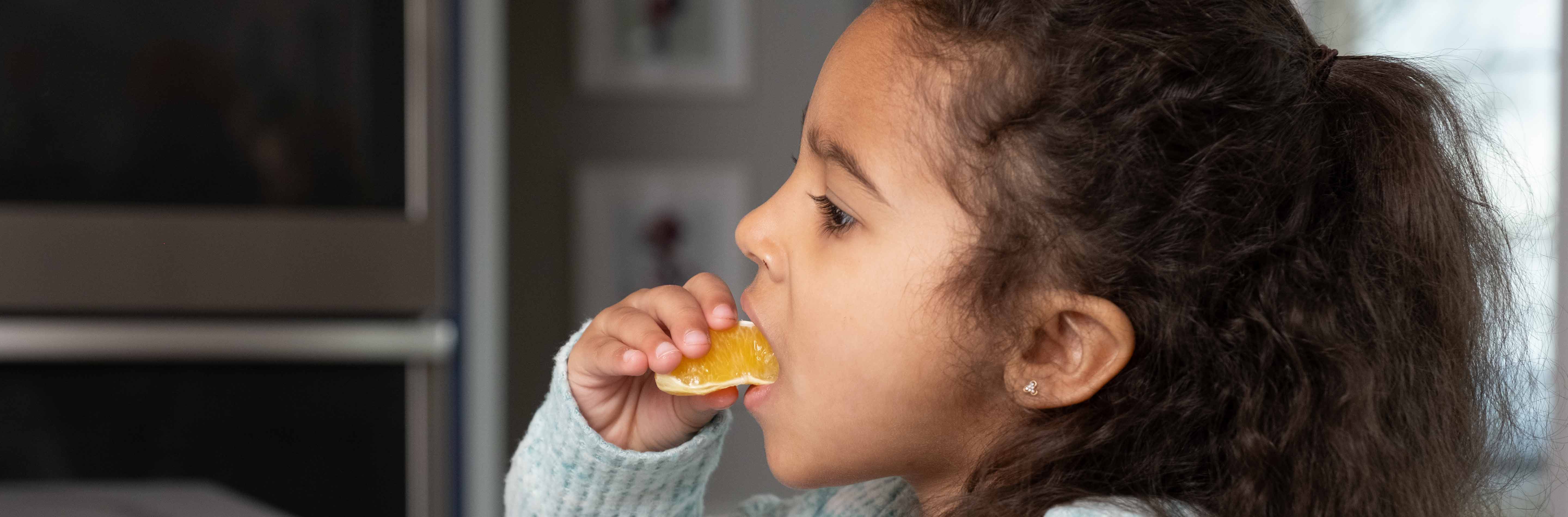 Toddler girl with curly brown hair sitting at table eating an orange.