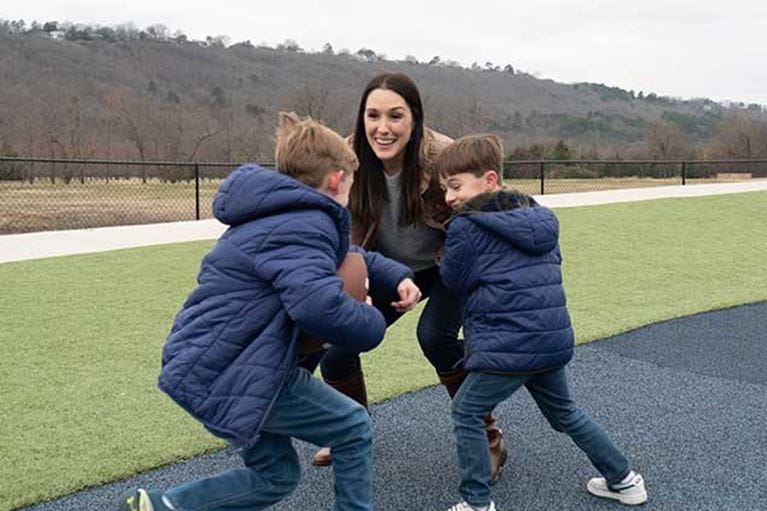 Mom playing football outside with sons in blue coats.