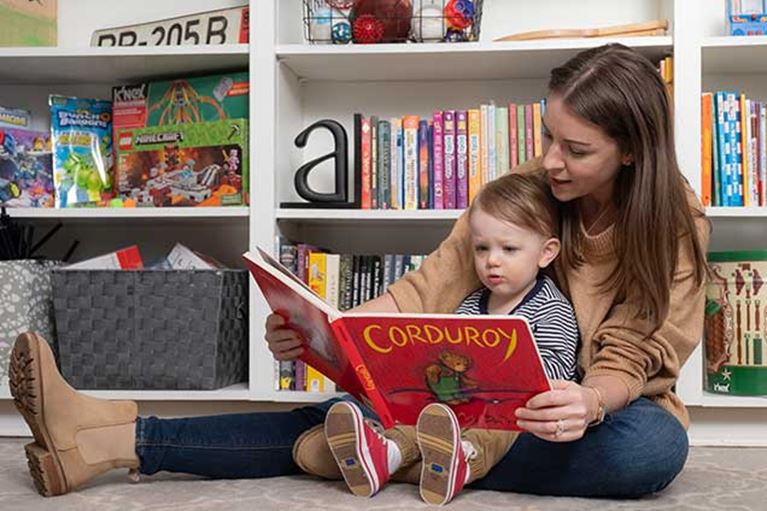 Mother sitting down reading to son in front of bookshelf.