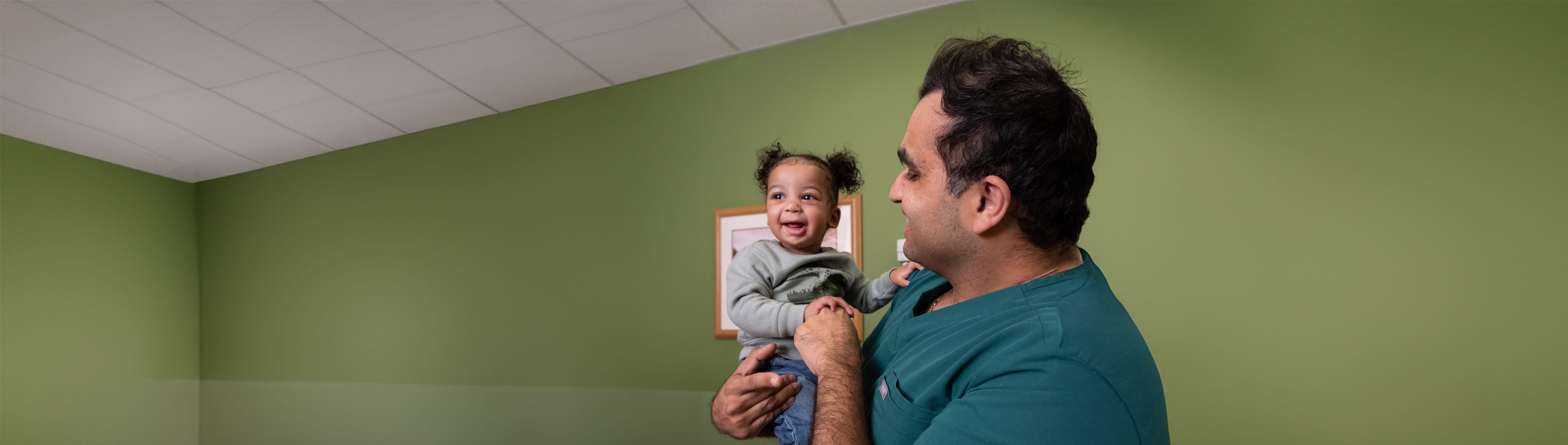 Baby boy with pigtails being held by doctor in green scrubs.