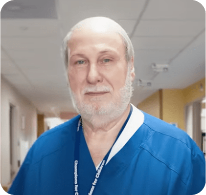 Male with beard standing in hallway wearing blue scrubs.