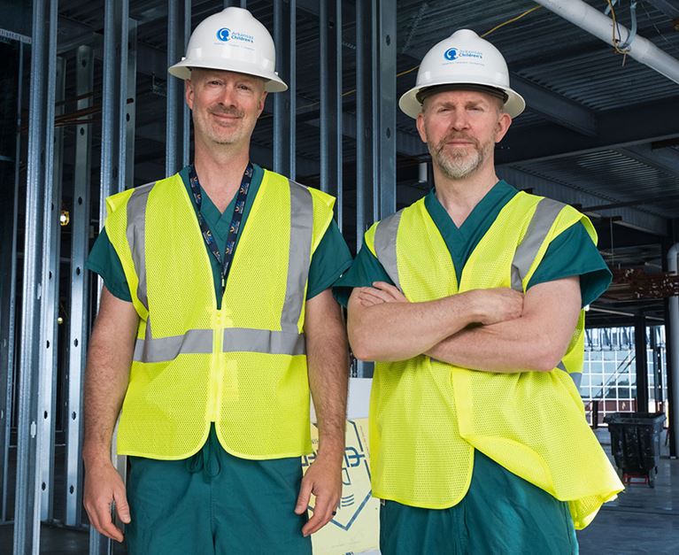 Two physicians wearing white hard hats standing in construction zone.