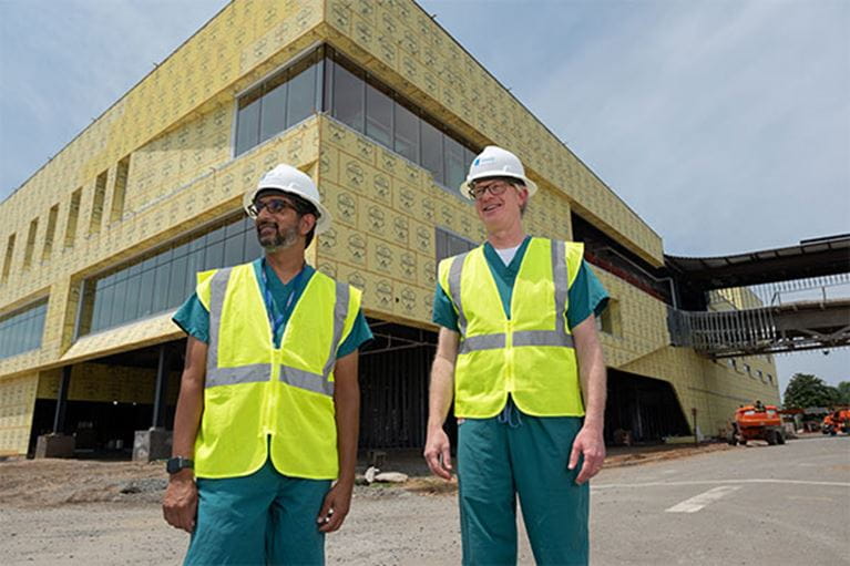 Two men wearing hard hats and safety vests outside of building being constructed.