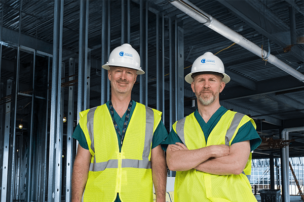 Two physicians wearing white hard hats standing in construction zone.
