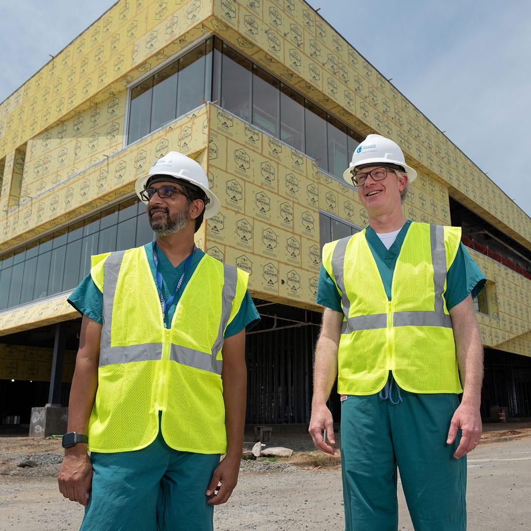 Two men wearing hard hats and safety vests outside of building being constructed.