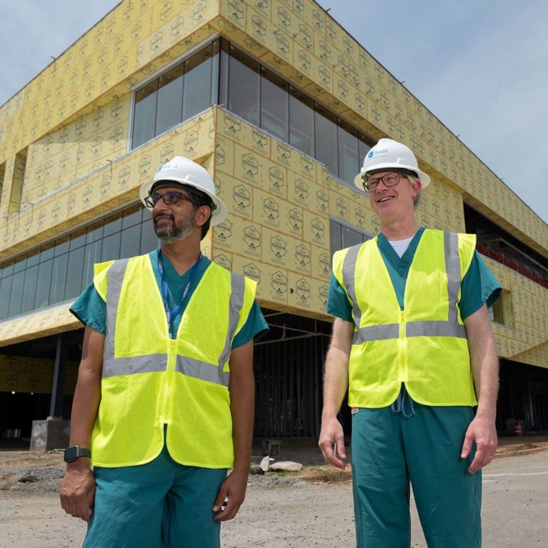 Two men wearing hard hats and safety vests outside of building being constructed.