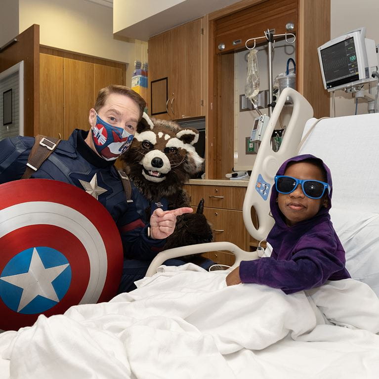 Captain America and Rocket Raccoon standing beside patient sitting up in hospital bed.