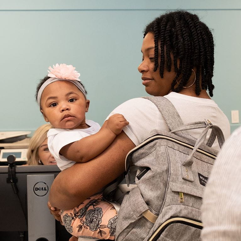 Girl wearing pink bow being held by her mother wearing backpack.
