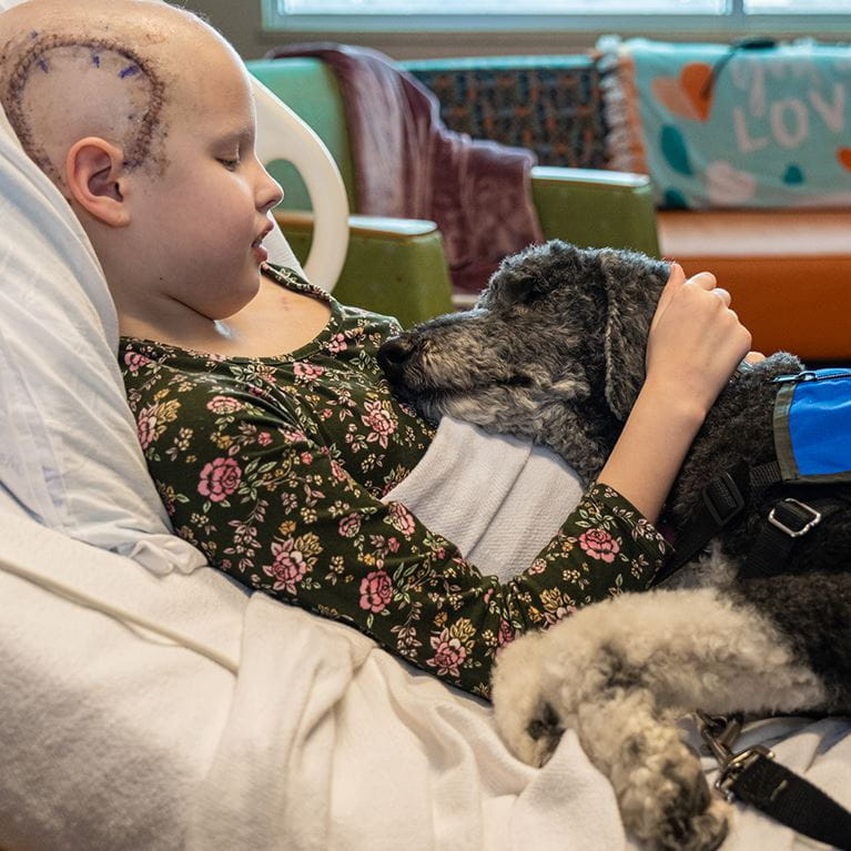Girl laying in hospital bed petting black dog.