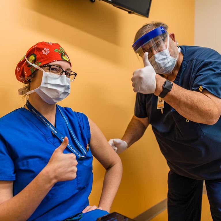 Nurse wearing scrubs and mask getting vaccinated by another nurse wearing gloves and mask.