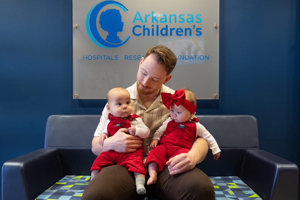 Dad holding twin babies wearing red overalls.